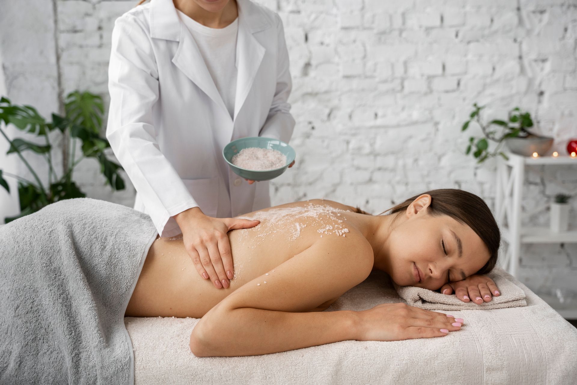 Woman receiving a back scrub at a spa. Therapist applying exfoliant, white brick wall background.