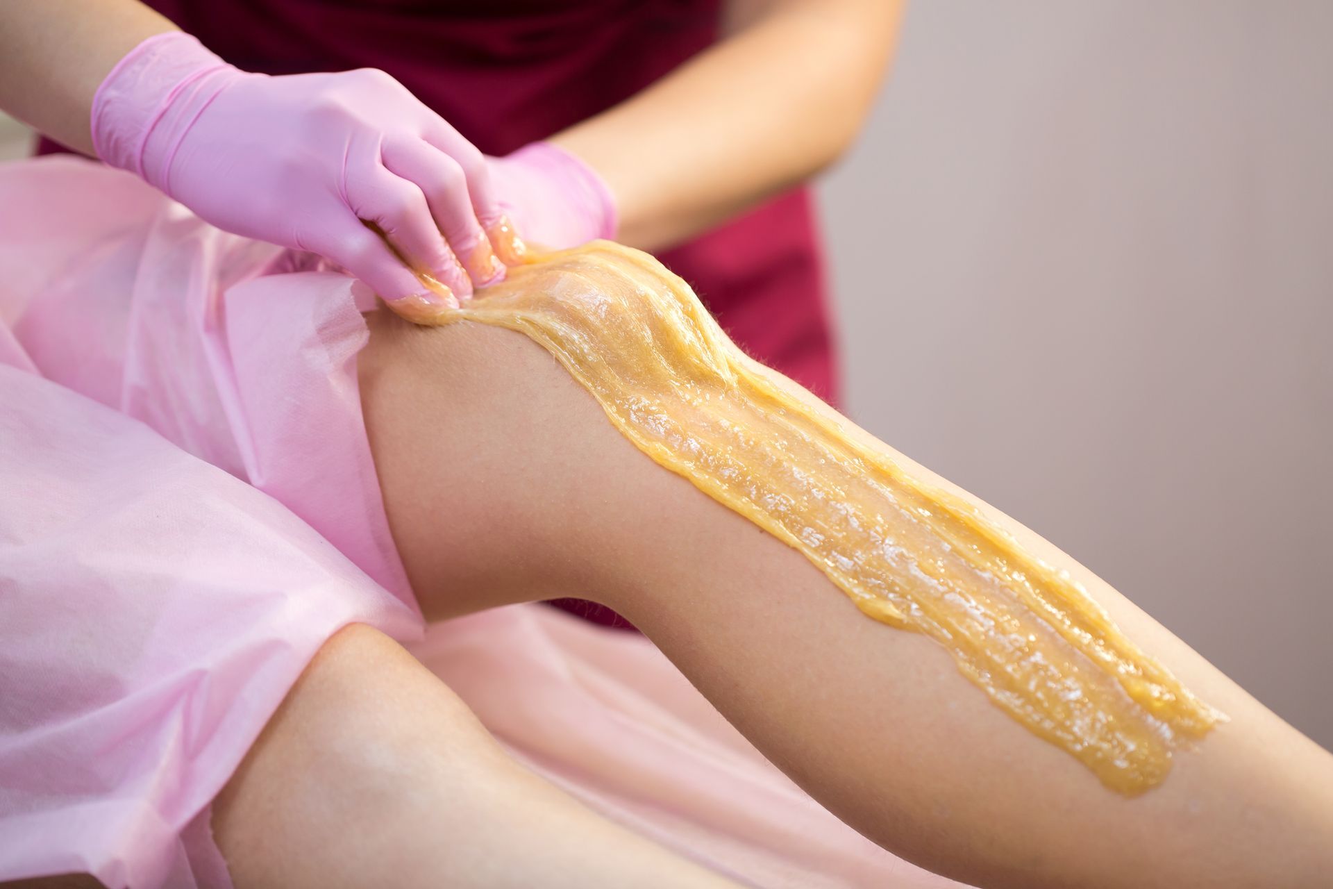 Leg being waxed. Esthetician in gloves applies warm wax to leg with a pink sheet covering surface.