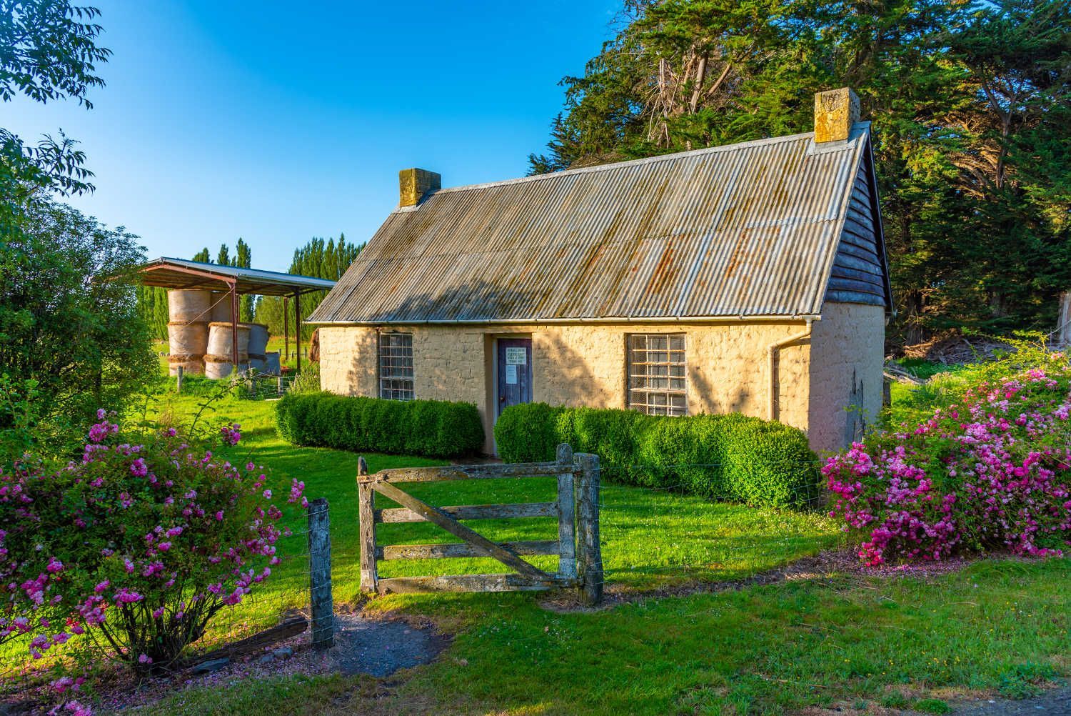 An old stone house with a wooden fence in front of it is surrounded by flowers.