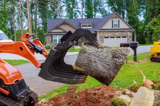 Orange excavator lifting a large tree stump in front of a suburban house and yard
