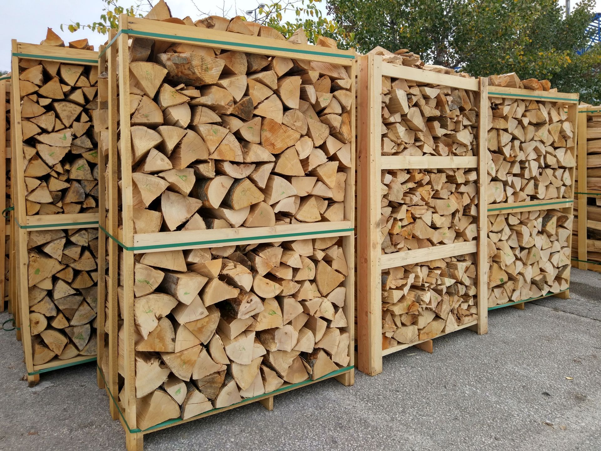 Stacked firewood in wooden racks outdoors on gravel, with tan split logs and green trim.