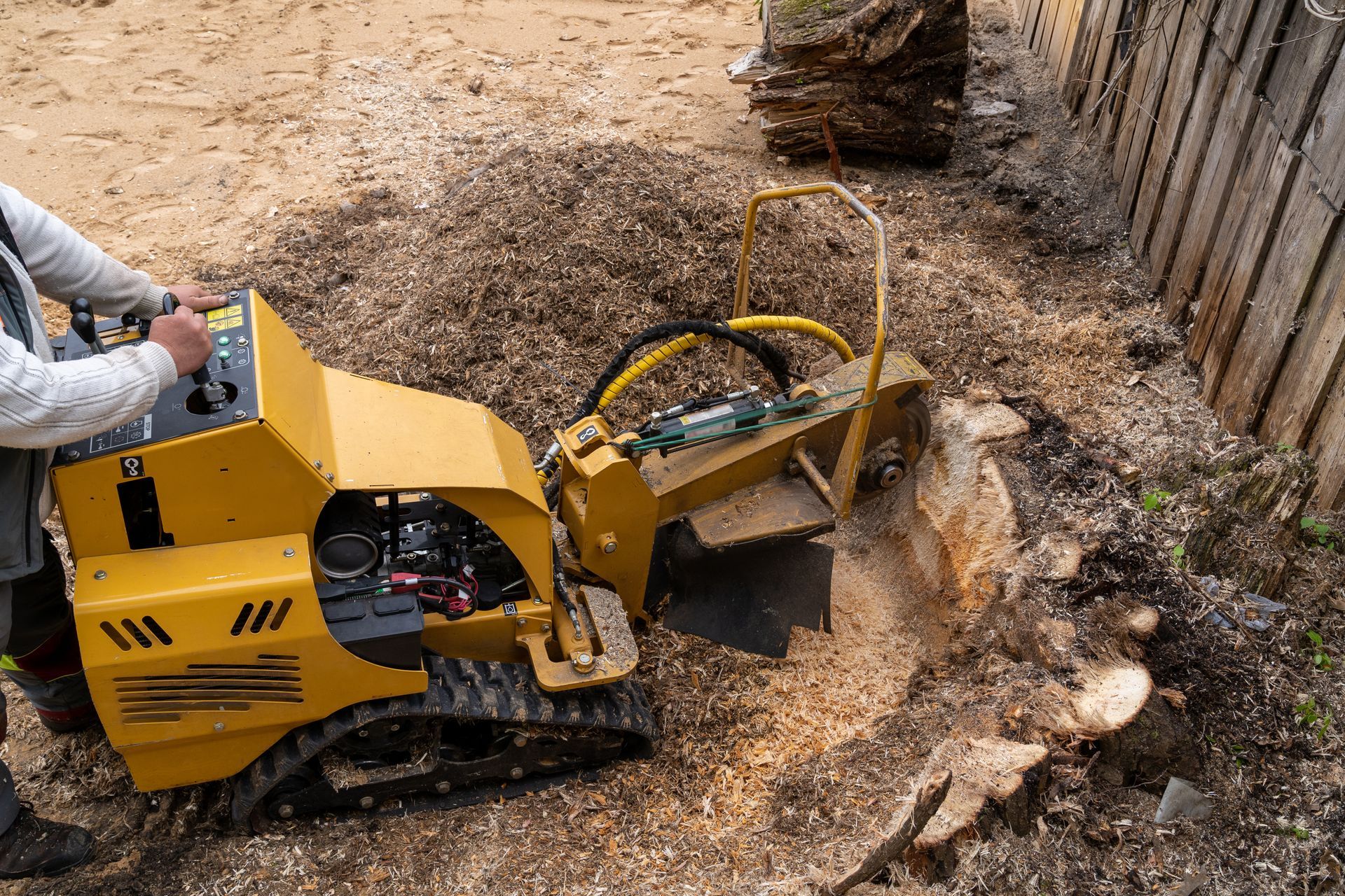 Yellow wood chipper clearing debris beside a fence, with a worker operating it outdoors.