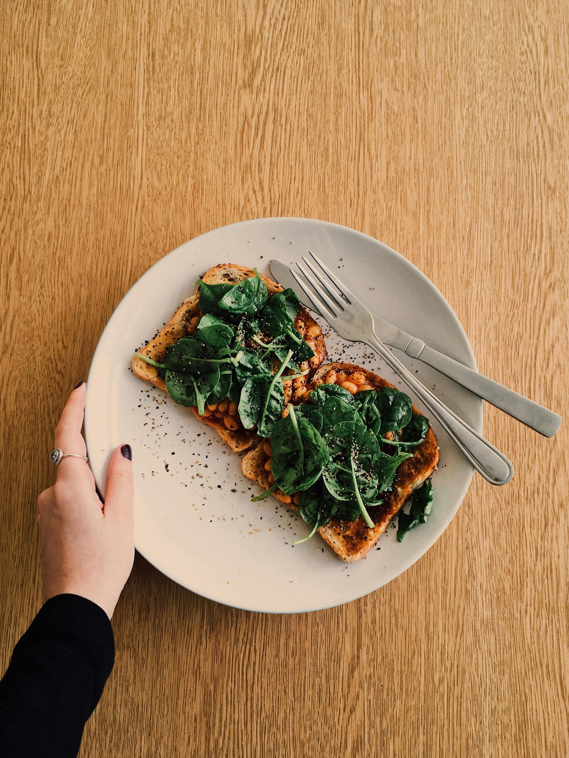 Toast topped with spinach on a white plate, hand holding the plate on a wooden table. Silverware is beside it — All Body Holistic Health in Thuringowa Central, QLD