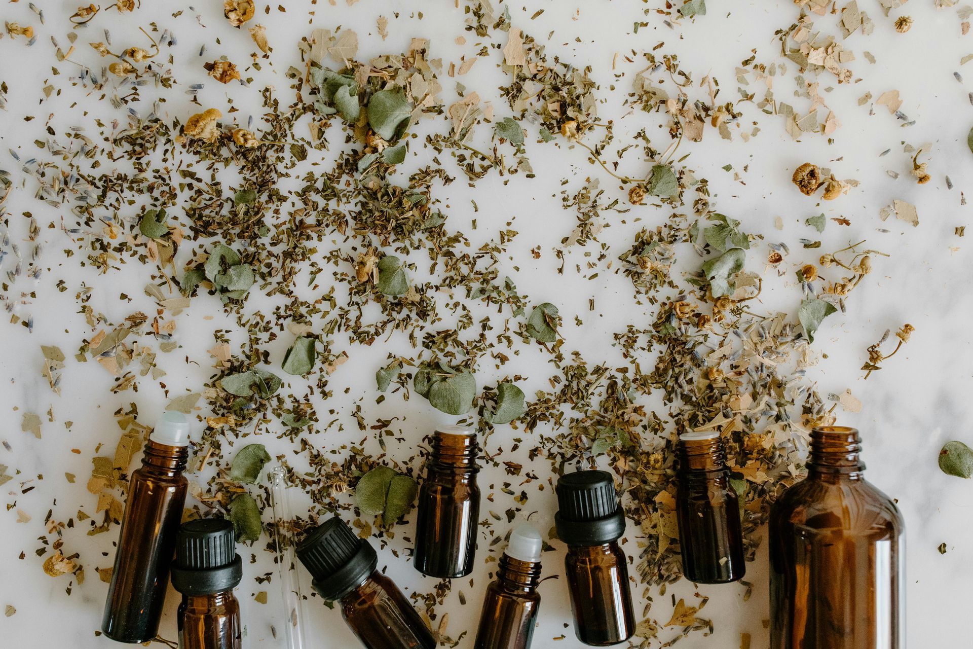 Brown glass essential oil bottles on a white surface scattered with dried herbs  — All Body Holistic Health in Thuringowa Central, QLD