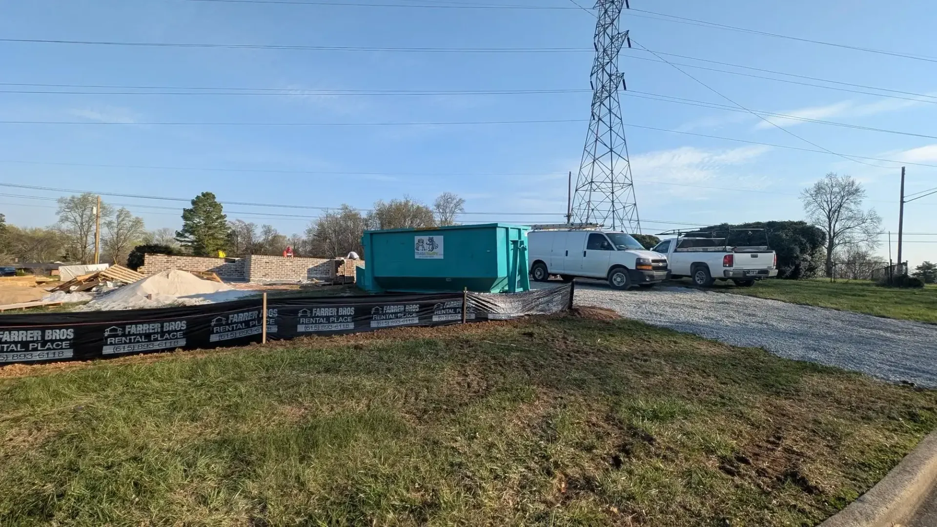 Construction site with a teal container, white van, gravel road, and utility tower under a clear sky