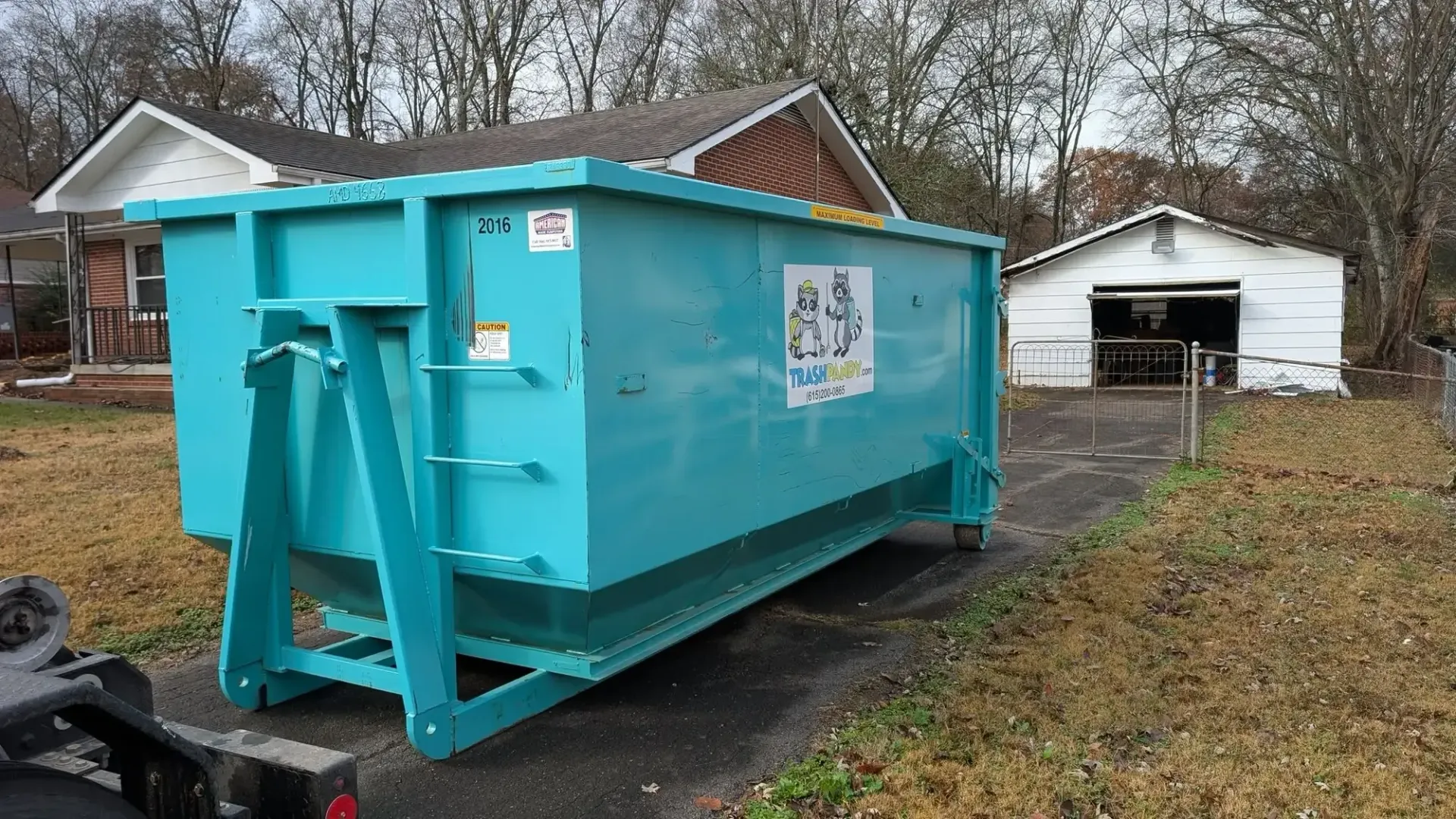 Bright turquoise dumpster on a trailer in a driveway, with houses and leafless trees in the background