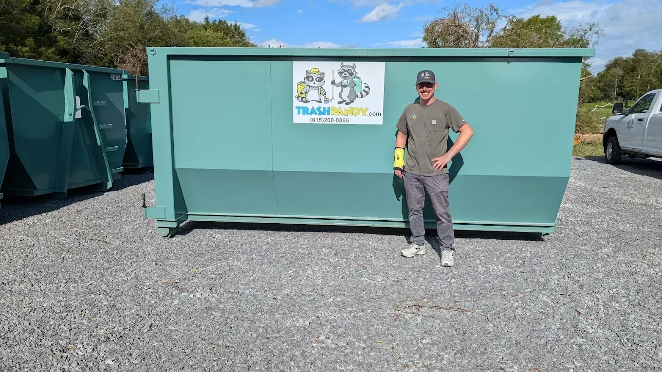 Man standing next to a large teal dumpster. Gravel ground, sunny day.