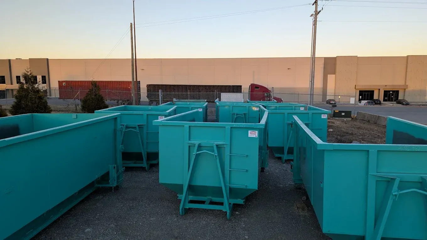 Several turquoise dumpsters in an outdoor lot, set against a warehouse and a sunset sky.