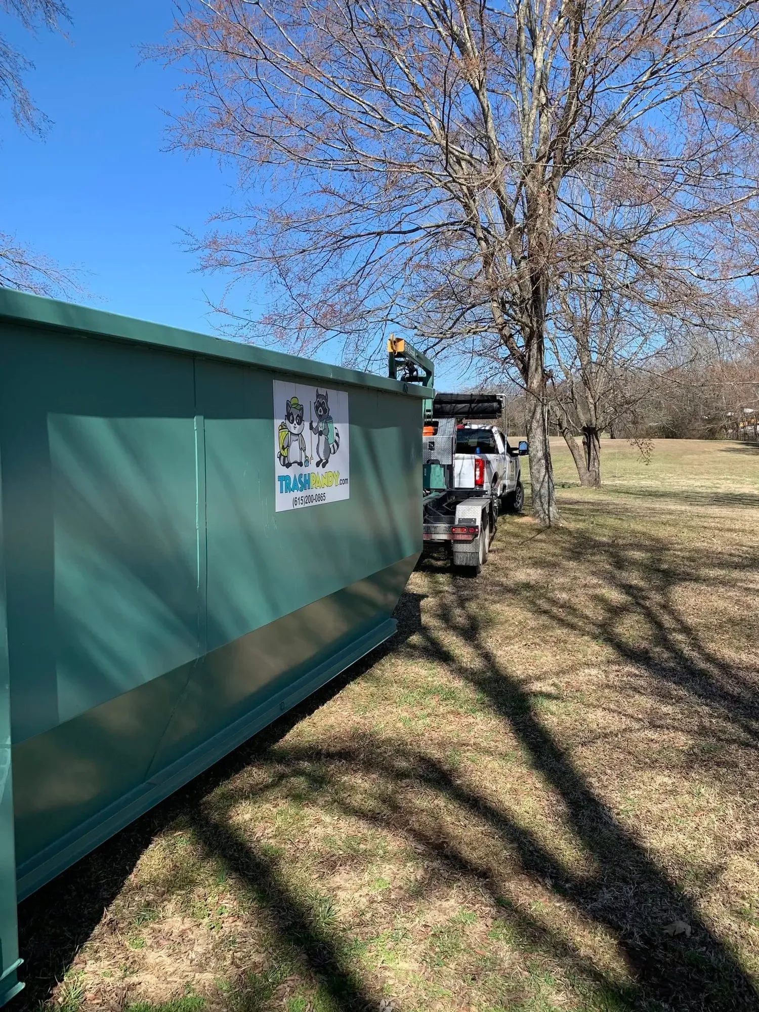 A large green dumpster next to a white pickup truck in a grassy area with a tree and blue sky.