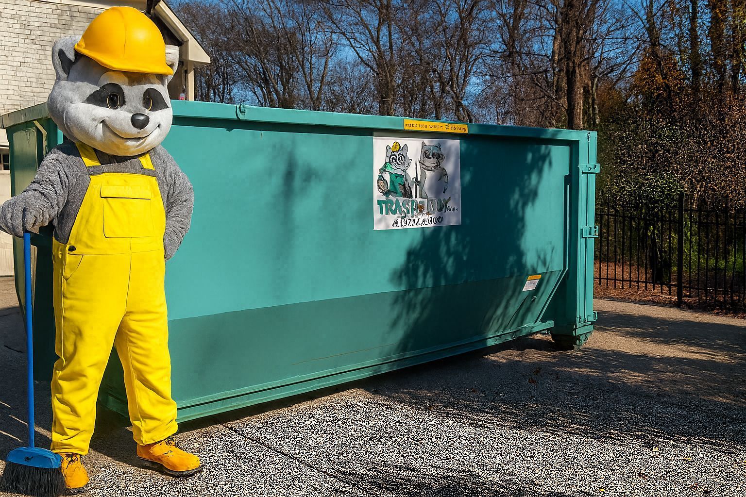 A raccoon mascot in yellow overalls and hard hat leans on a broom next to a turquoise dumpster.