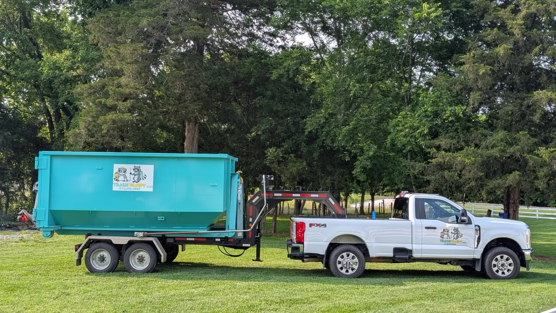 White truck towing a teal dumpster on a trailer, parked in grassy field.