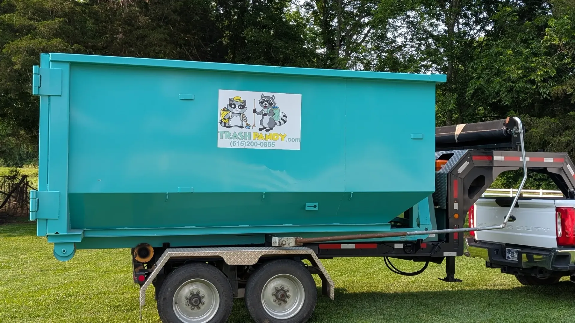 A teal dumpster on a trailer, hitched to a white pickup truck, with a logo of two raccoons.