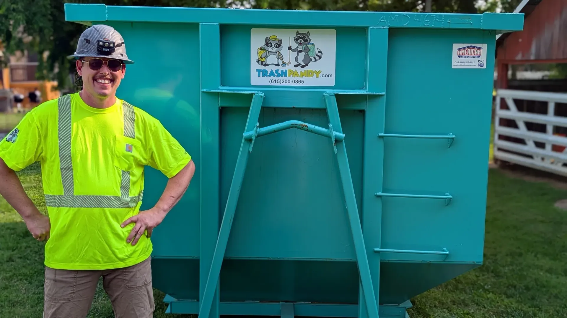A green dumpster next to a white truck on a grassy area under a tree on a sunny day.