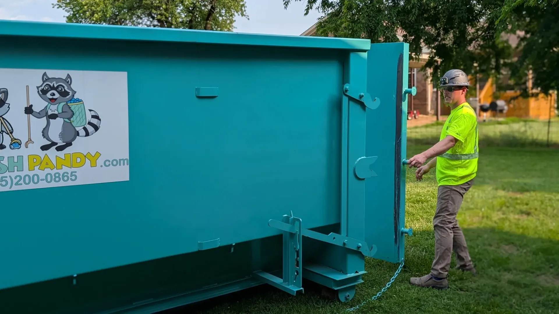 A man is standing next to a blue dumpster in a grassy field.