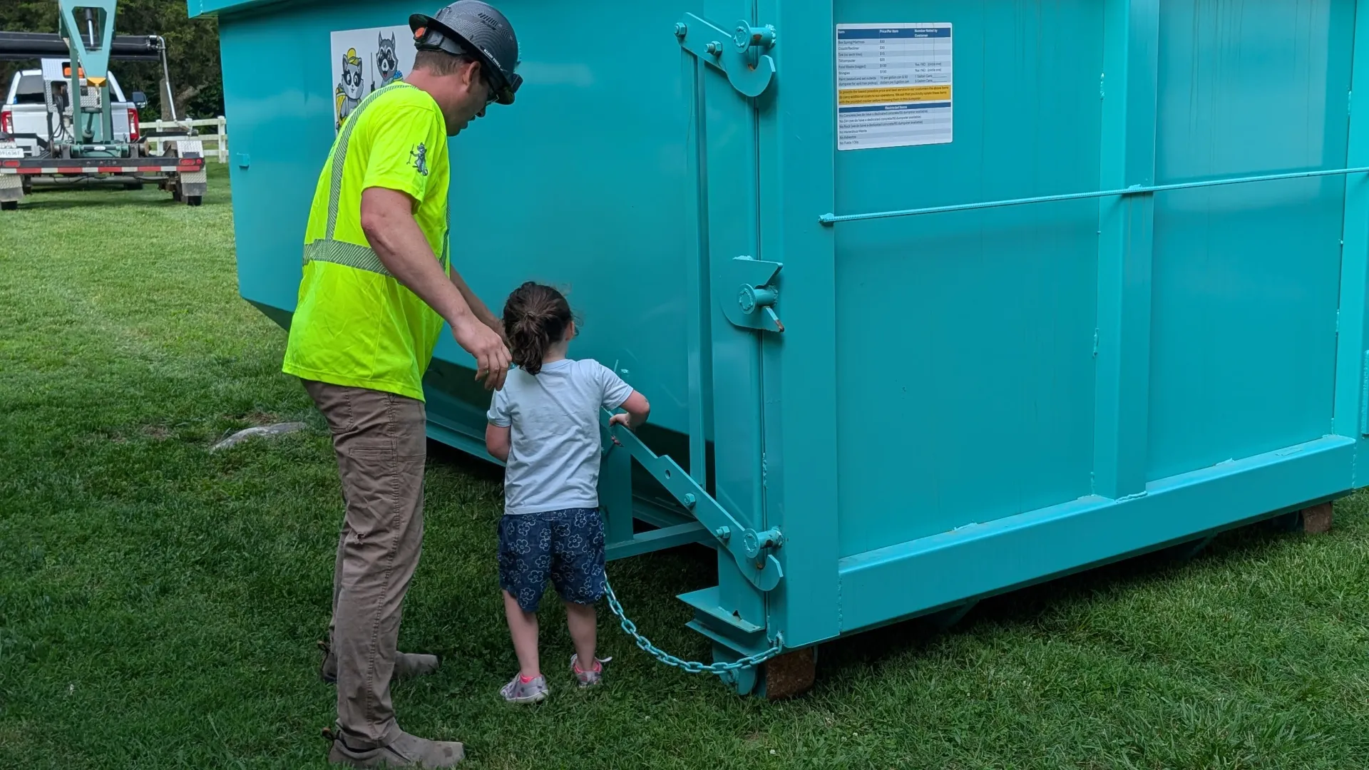Man in neon vest helps child open a large teal dumpster outdoors.
