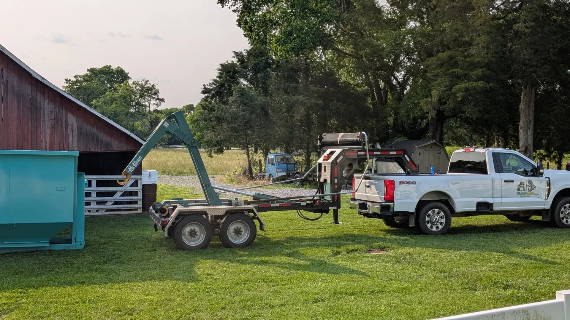 A white truck pulling a trailer with a lift is parked near a teal dumpster and a red barn.