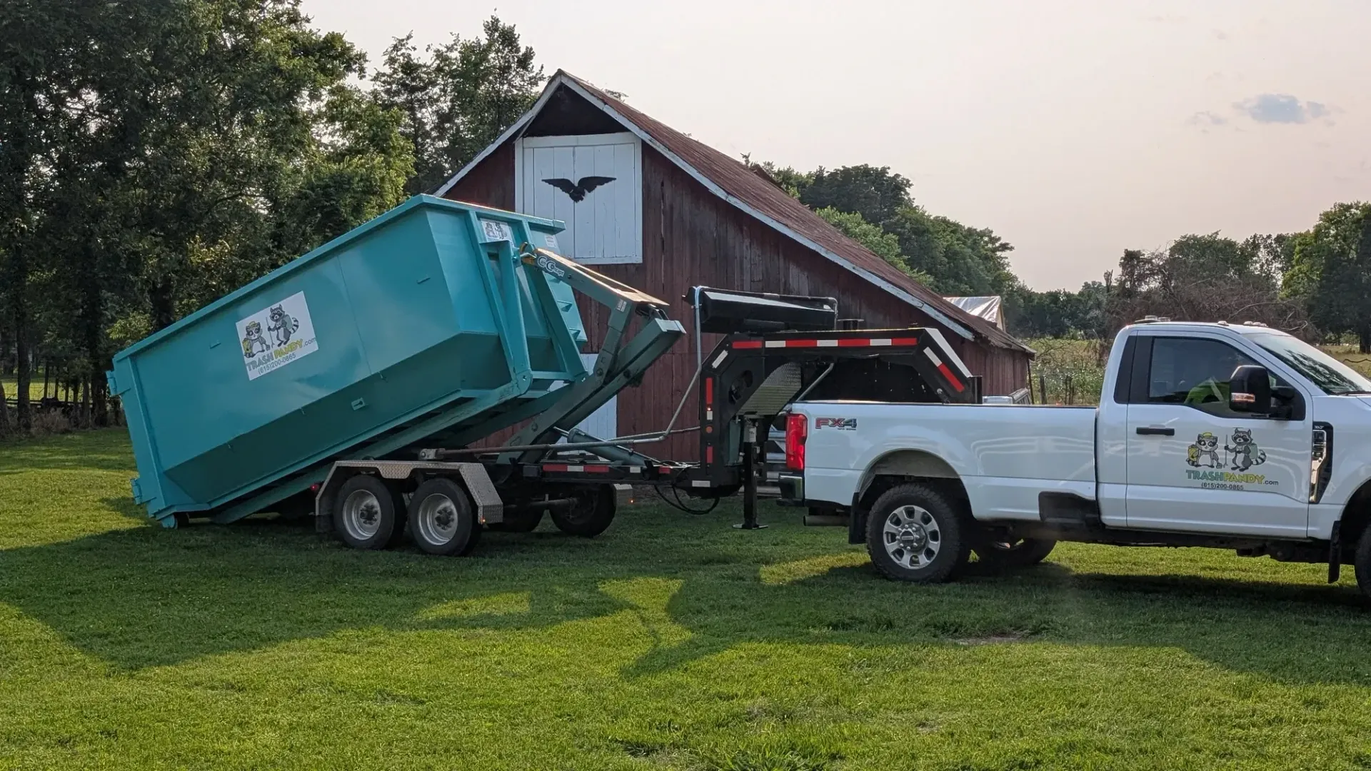 A white pickup truck towing a teal dumpster trailer on a grassy field in front of a red barn.