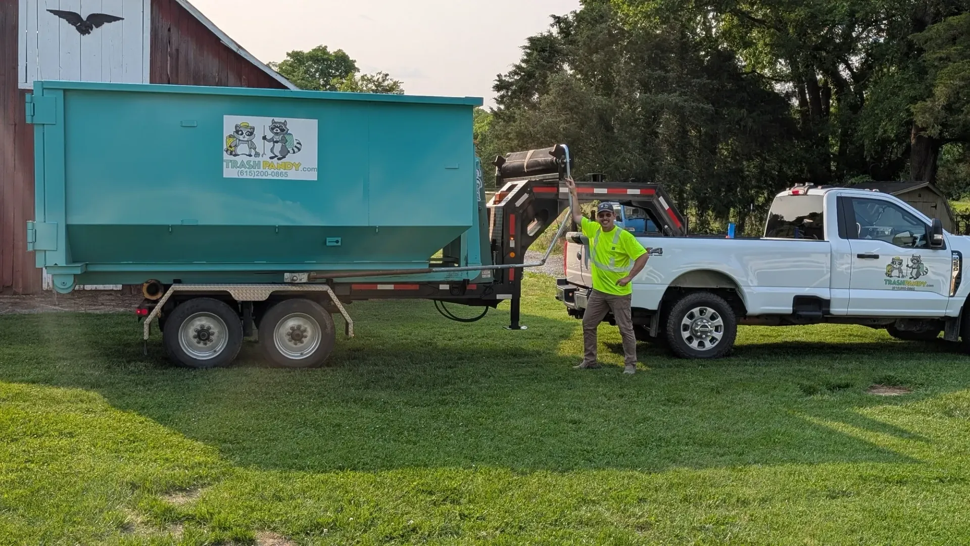 Man in safety vest near a truck towing a teal dumpster trailer on a grassy area in front of a barn.
