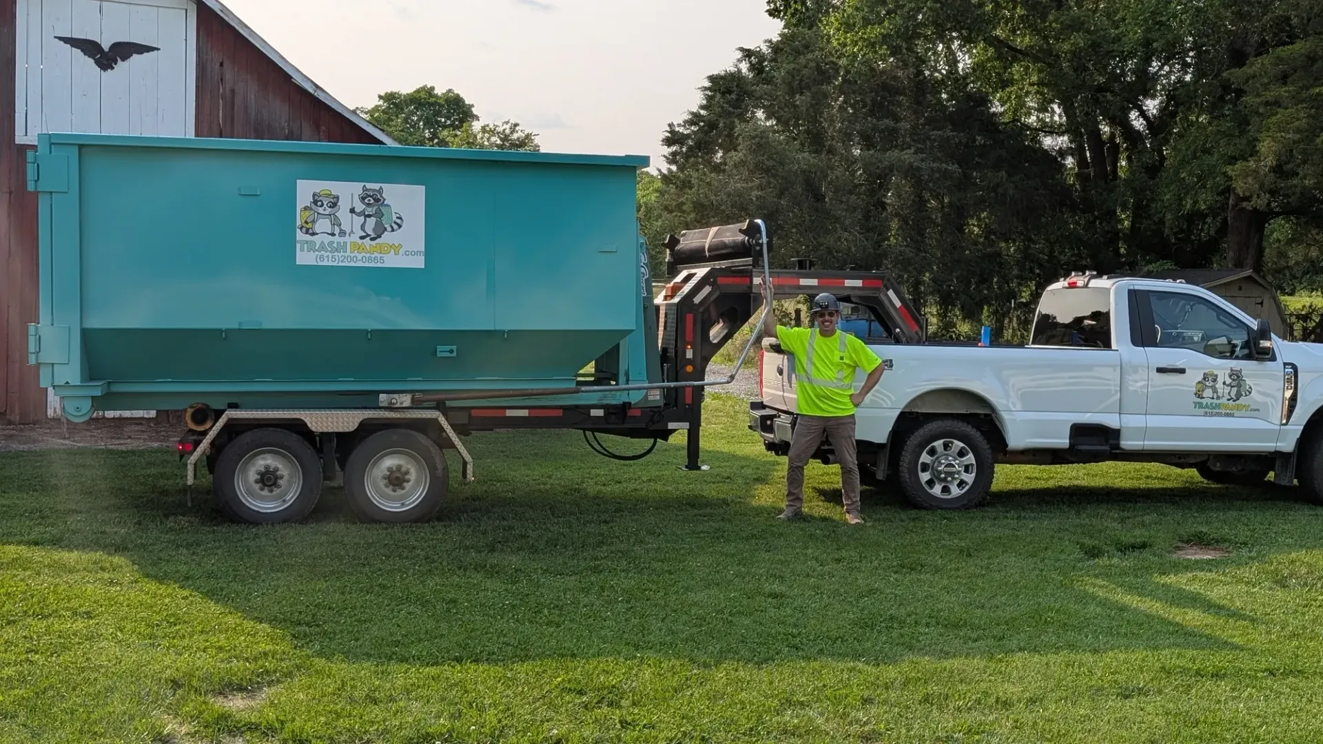 A person stands next to a white truck with a green dumpster on a trailer in front of a barn.