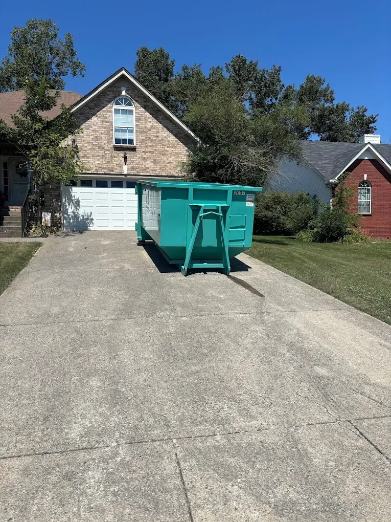 Green dumpster in a driveway in front of a house.