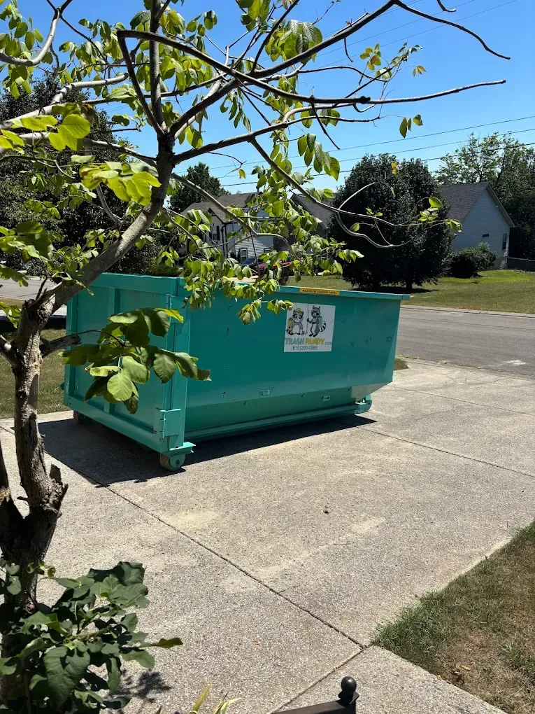 A large teal dumpster sits on a driveway in front of a suburban home on a sunny day.