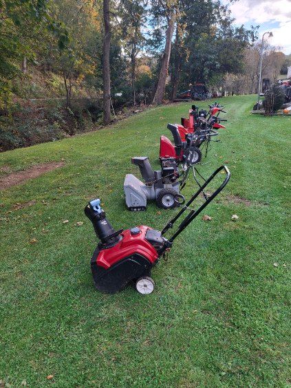 Snowblowers lined up on a grassy lawn. Red and gray machines face the viewer, a creek and trees in the background.