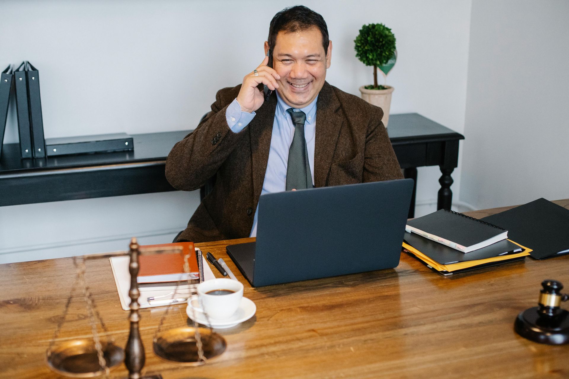 Abogado de traje hablando por teléfono, sonriendo frente a una computadora portátil, sentado en un escritorio.