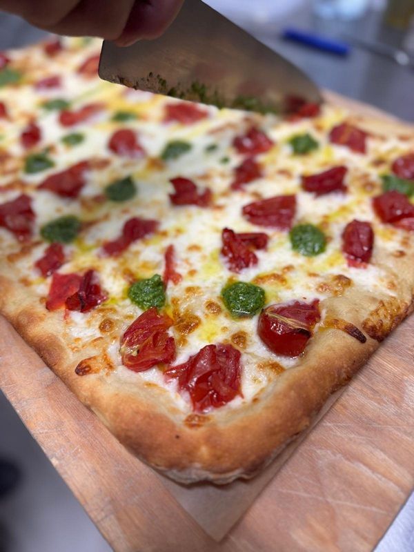 Pizza quadrata con peperoni e salame piccante su un tagliere di legno, tagliata con un coltello da pizza.