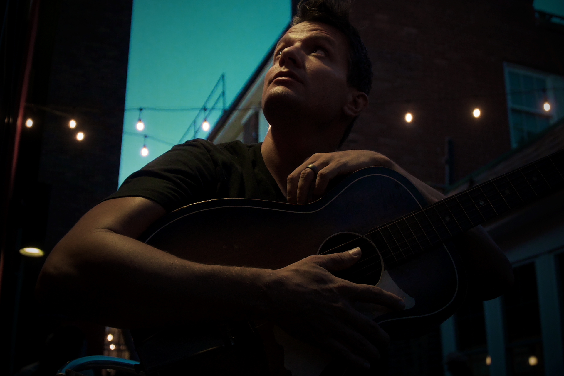 A man is playing an acoustic guitar in a dark alleyway.