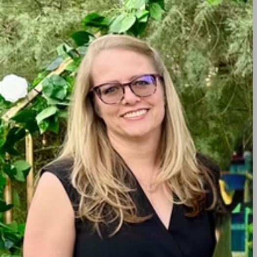 Tanya Paddock of Tanya Lynn Weddings wearing glasses smiles outdoors, in front of greenery.