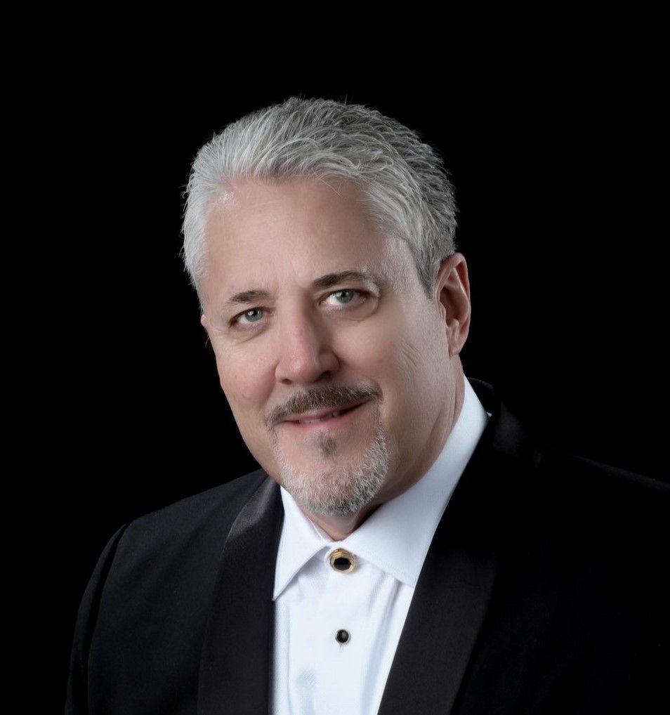 A silver-haired man with a mustache is wearing a white shirt and black jacket