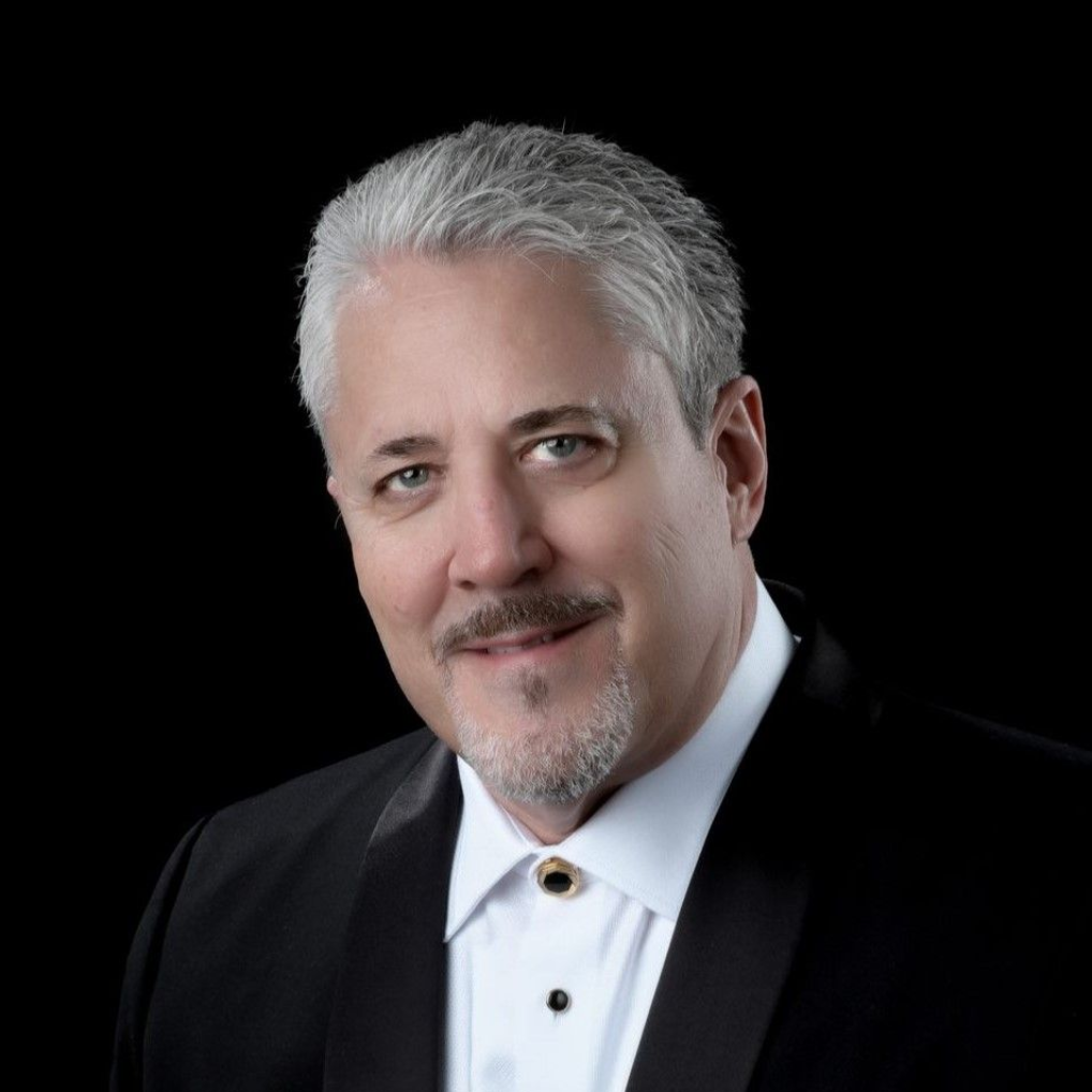 A silver-haired man with a mustache is wearing a white shirt and black jacket