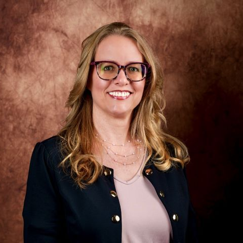 Tanya Paddock of Tanya Lynn Weddings wearing glasses smiles outdoors, in front of greenery.