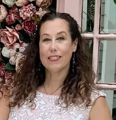 A woman in a pink dress is standing in front of a wall of flowers.