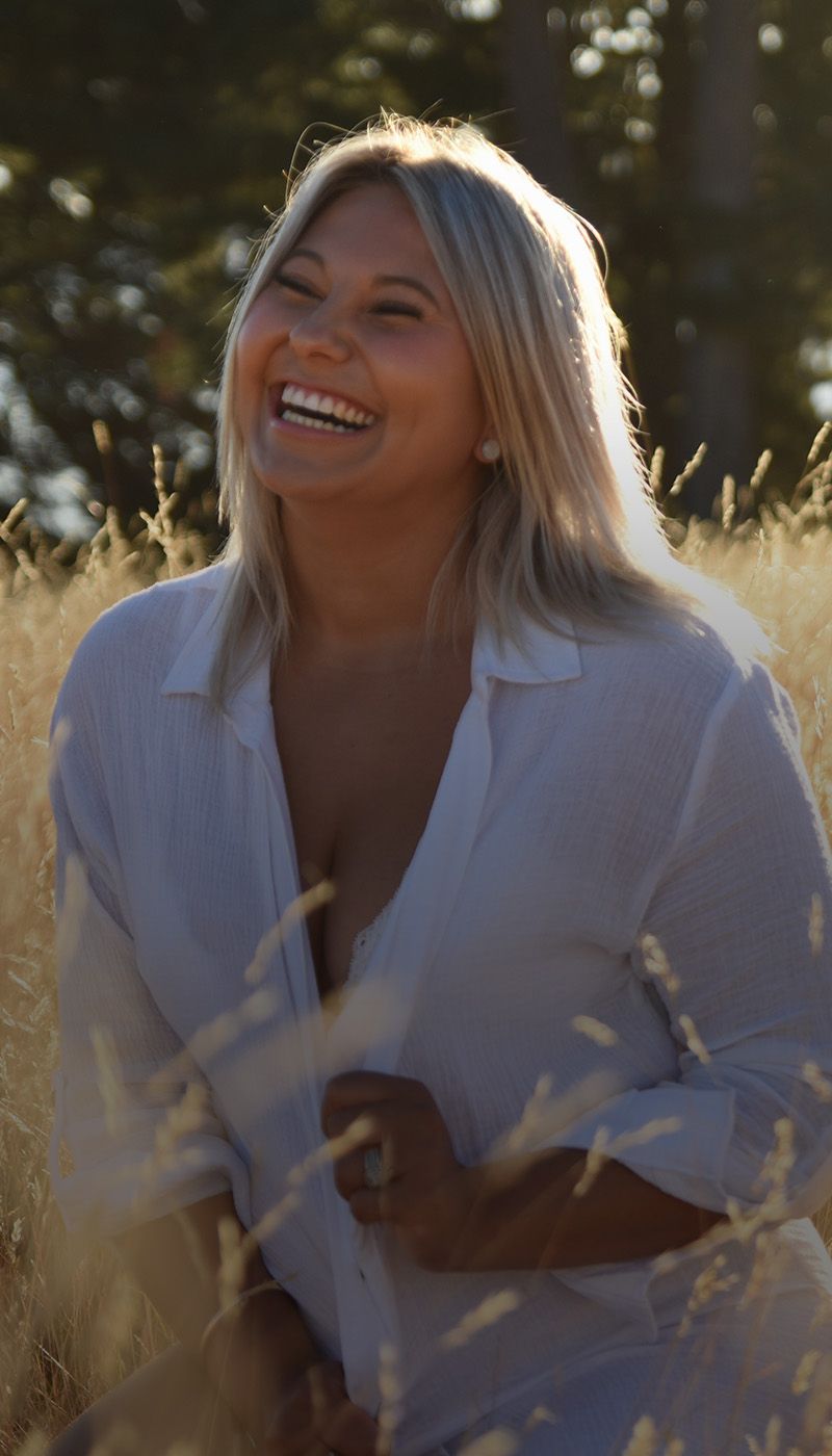 A woman in a white shirt is sitting in a field of tall grass and smiling.