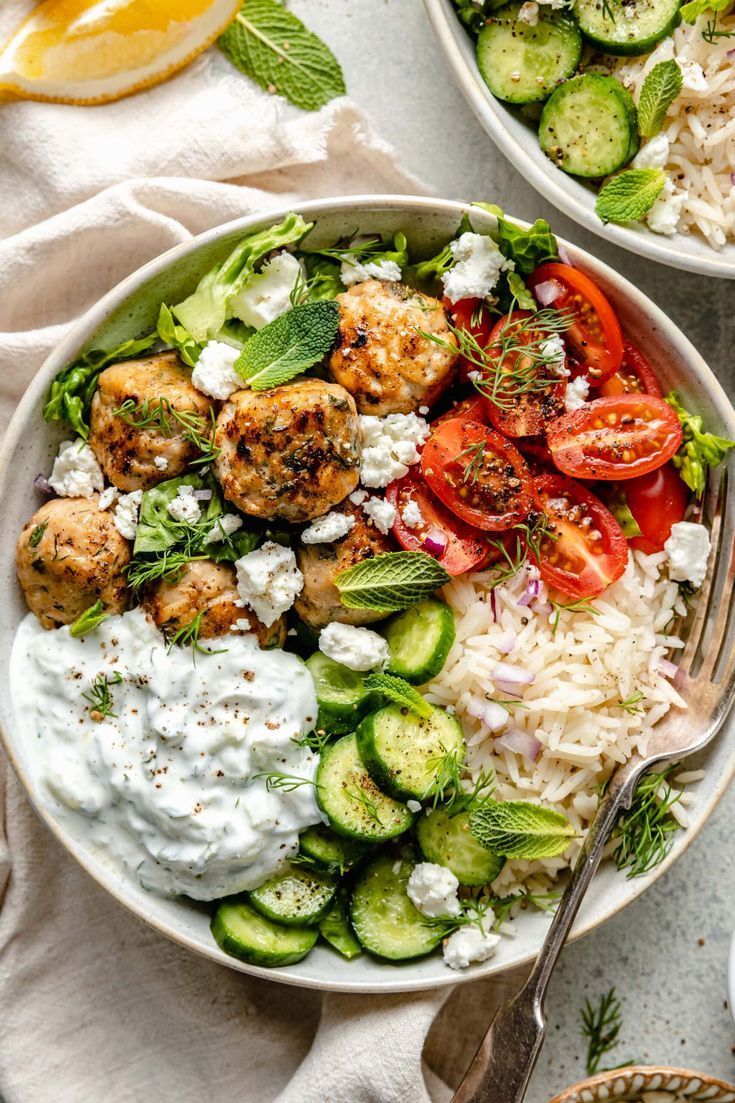 A bowl of food with meatballs , rice , tomatoes , cucumbers and yogurt sauce.