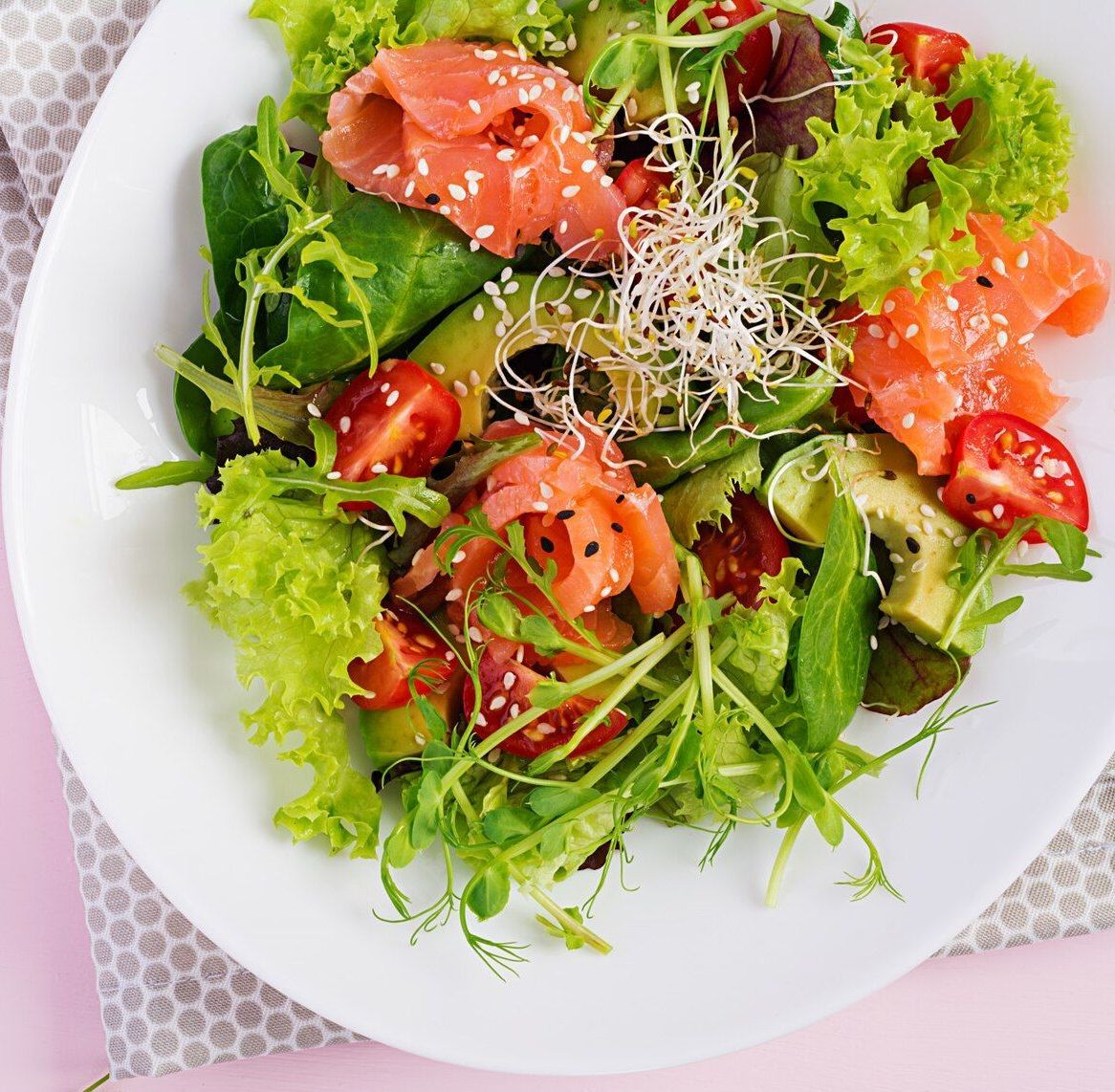 A white plate topped with a salad with salmon , tomatoes , lettuce and sesame seeds.