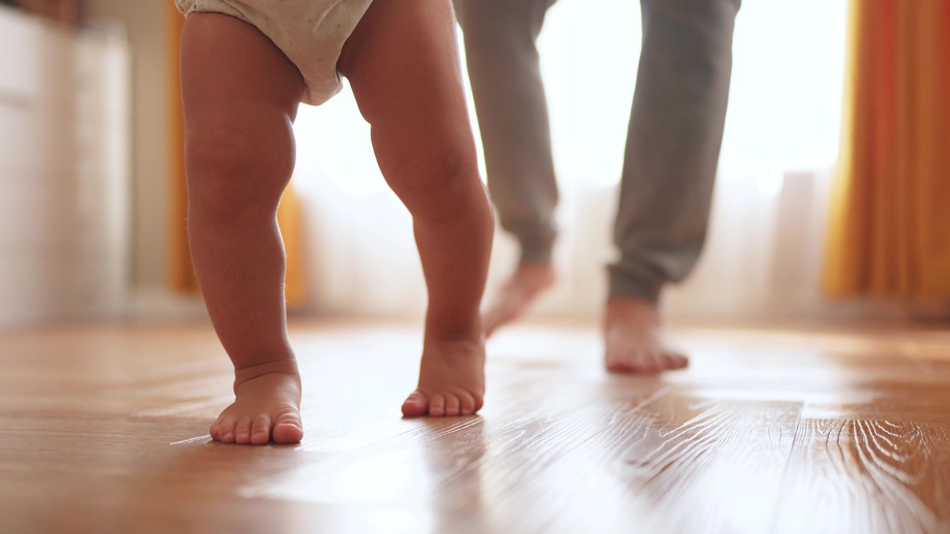 A woman and a baby are walking barefoot on a wooden floor.