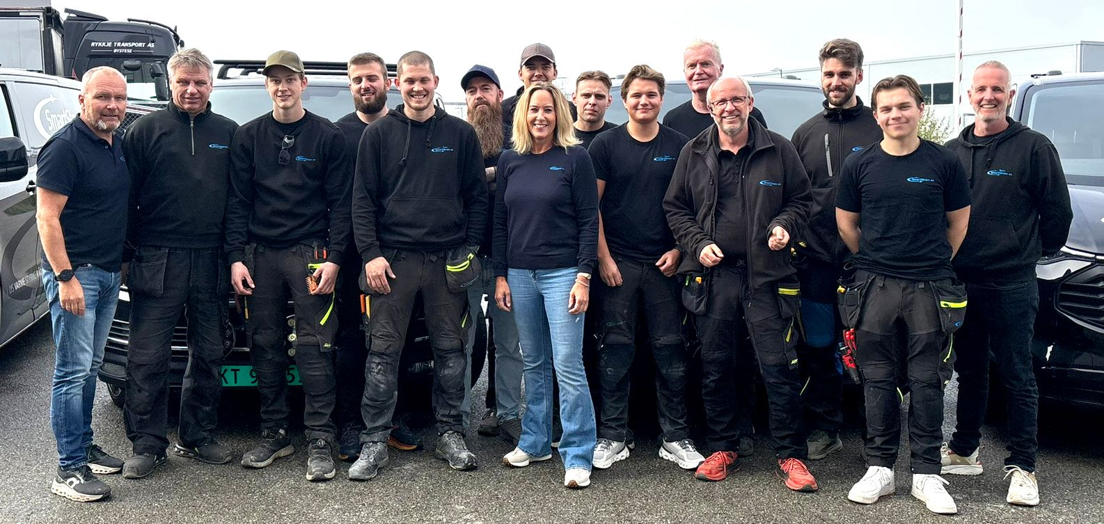 En stor gruppe mennesker i fritidsklær står sammen for et bilde på en utendørs parkeringsplass i nærheten av flere parkerte kjøretøy.