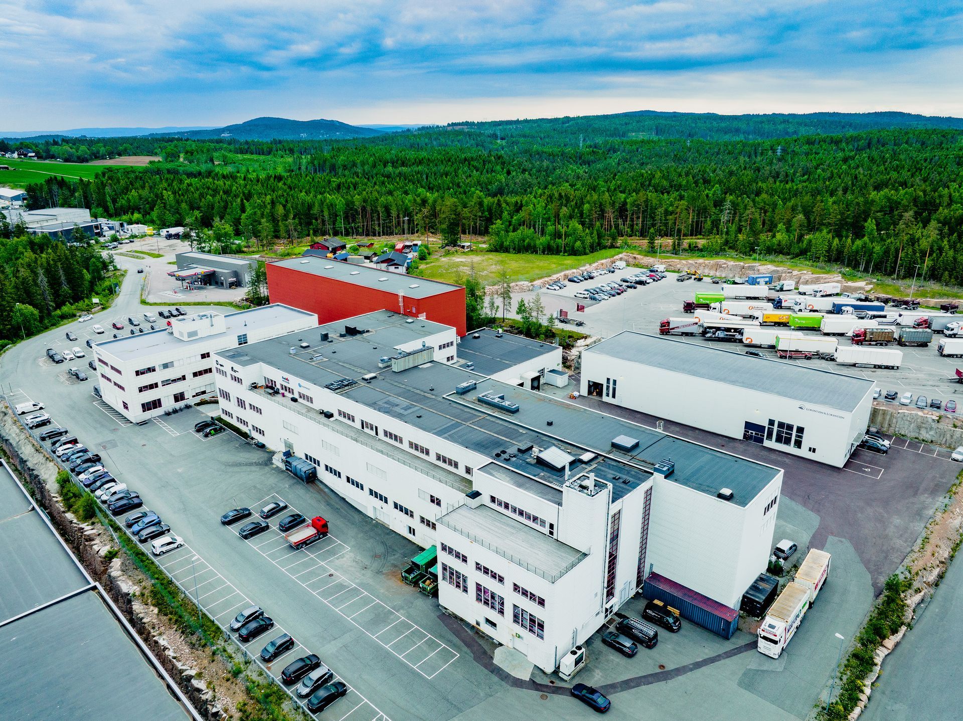Luftfoto av et stort hvitt kommersielt bygningskompleks med rød takseksjon, parkeringsplasser og lastebiler, nær en skog.