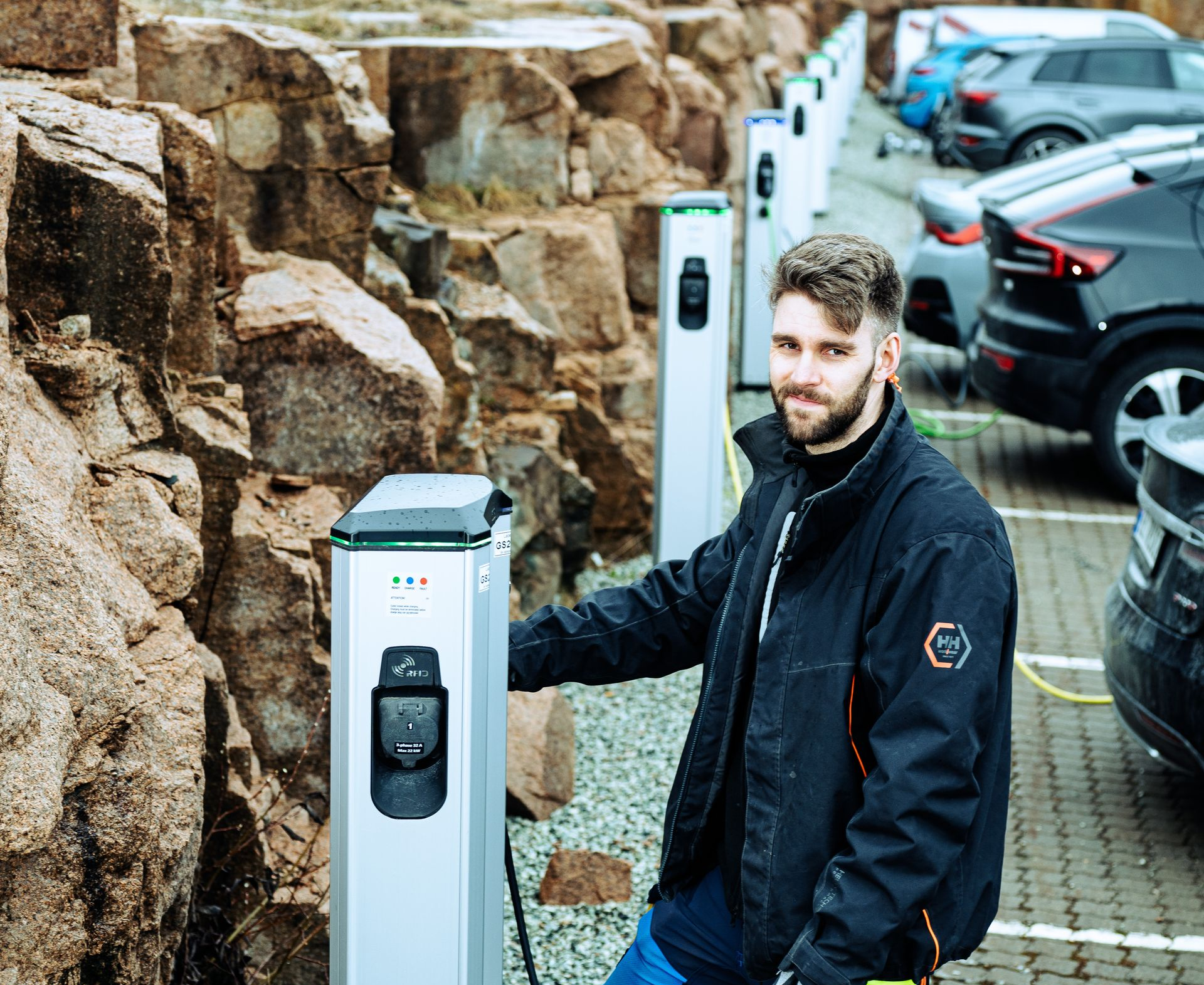 En person står på en parkeringsplass ved siden av en ladestasjon for elbiler, med steinete klipper i bakgrunnen.