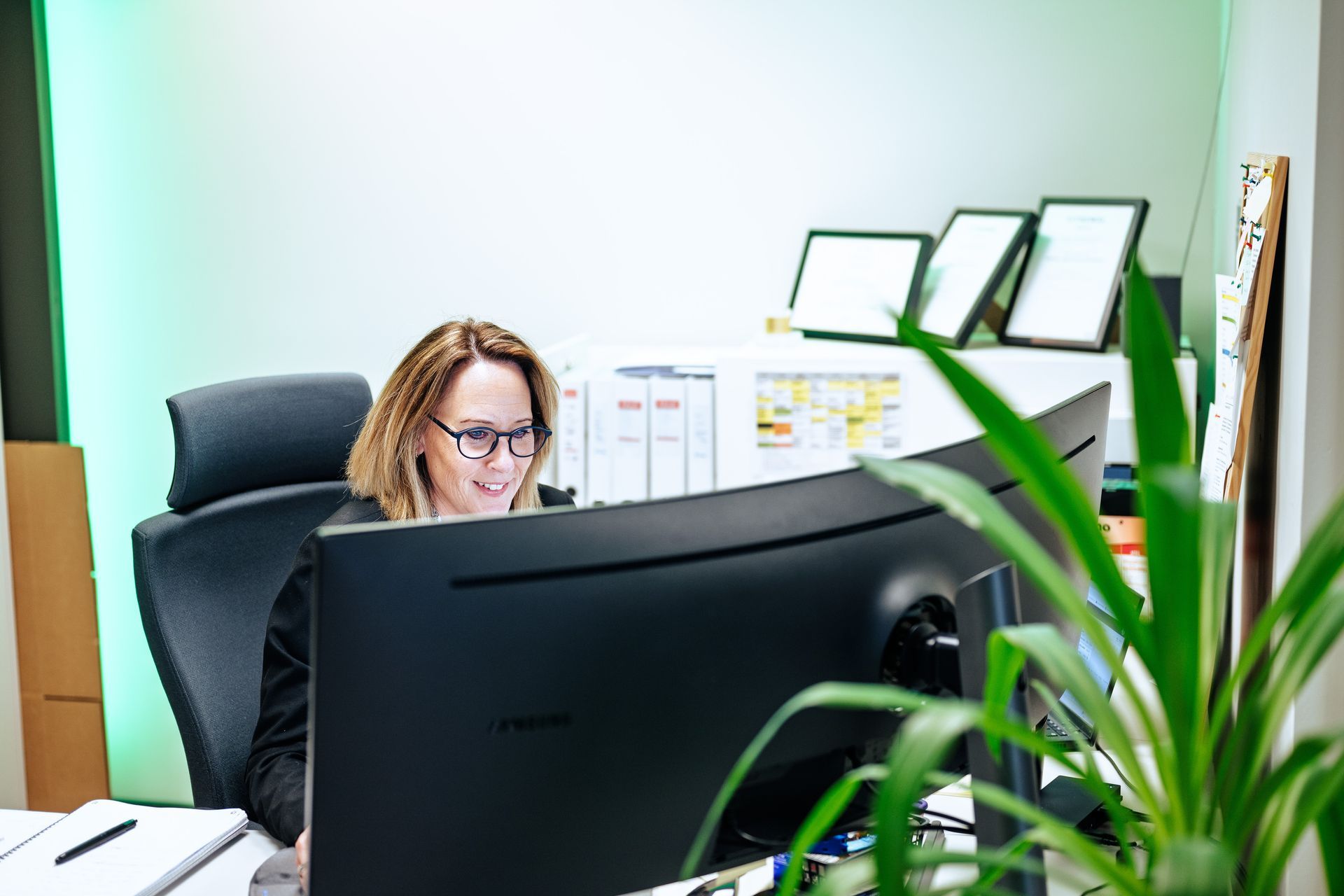 En person i et profesjonelt kontormiljø jobber ved et databord med flere skjermer og en plante i forgrunnen.