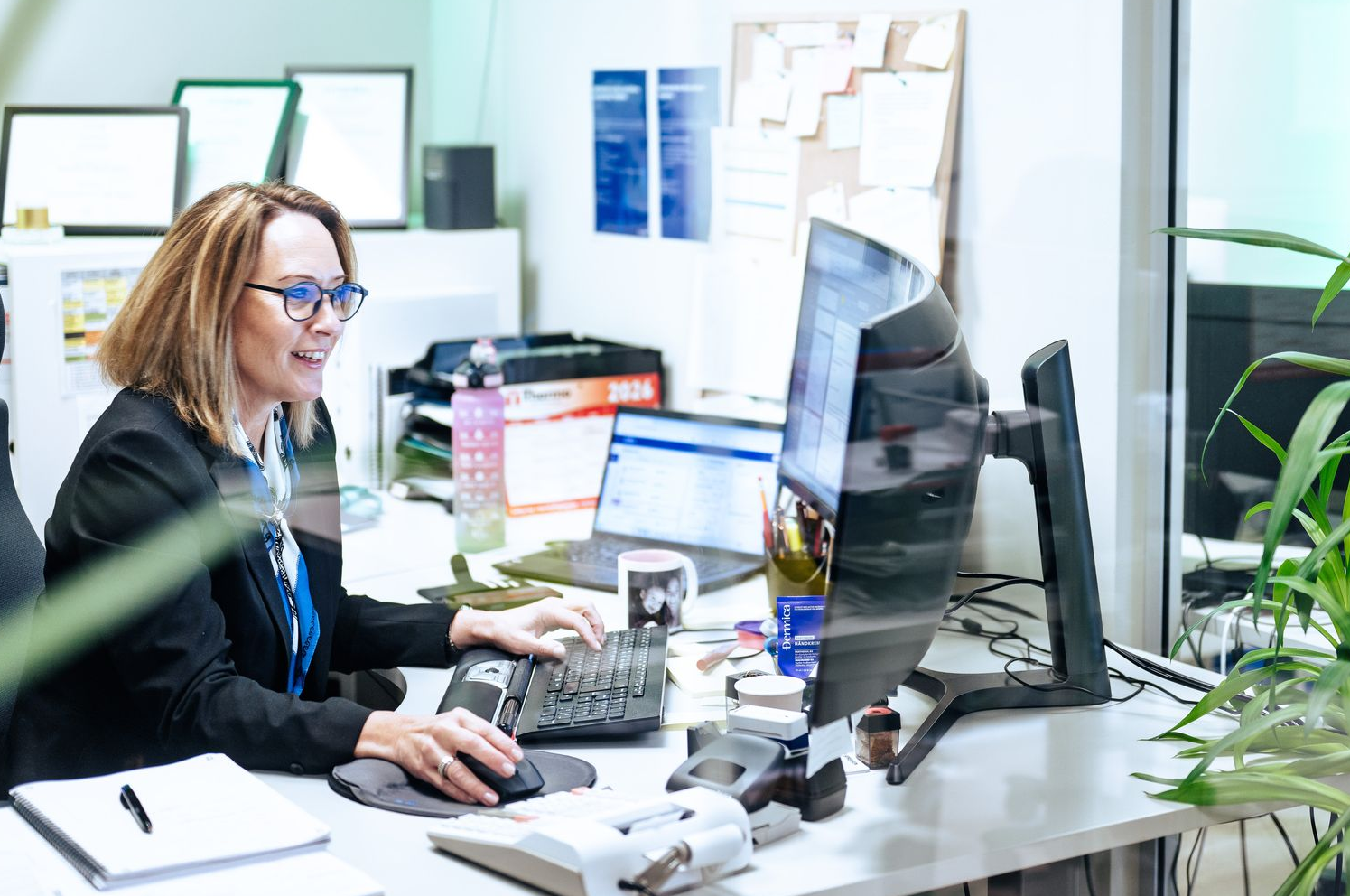 En person i et profesjonelt kontormiljø jobber ved et databord med flere skjermer og en plante i forgrunnen.