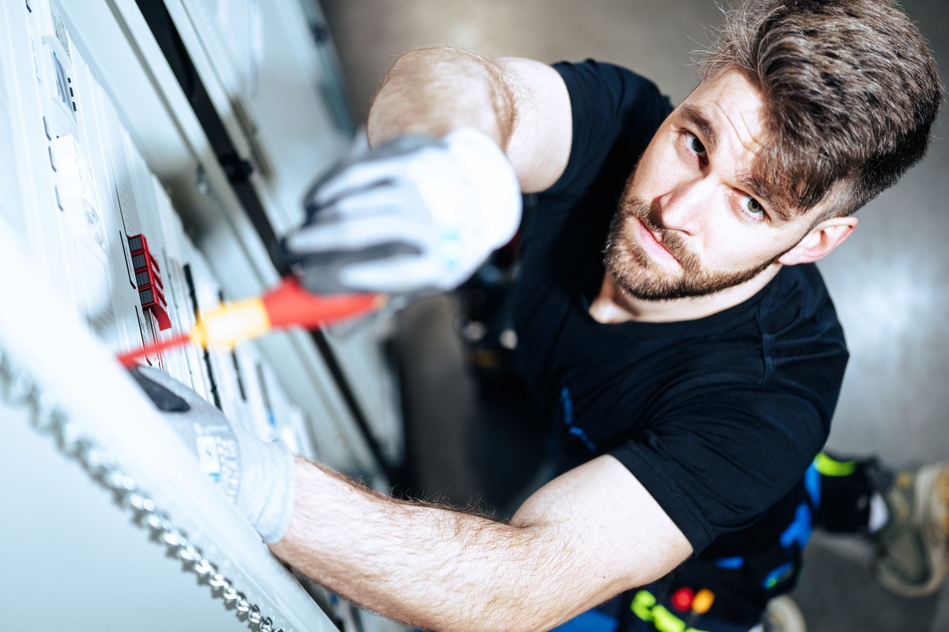 A person wearing work gloves uses a screwdriver to perform maintenance on a white industrial control panel.