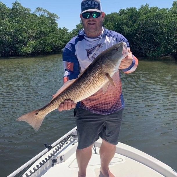 Man on boat holding a large redfish. Green trees in background. Blue sky, sunny day.