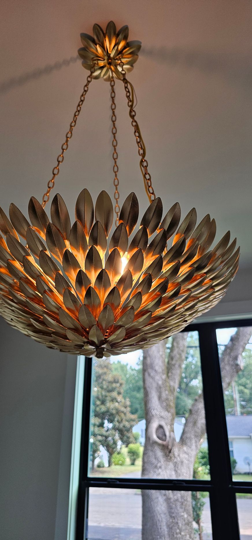 Golden chandelier shaped like a flower, hanging in front of a window with a tree and a view of a house in the background.