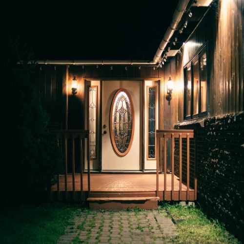 A dimly lit front porch at night. A white door with an oval glass window is framed by sidelights.