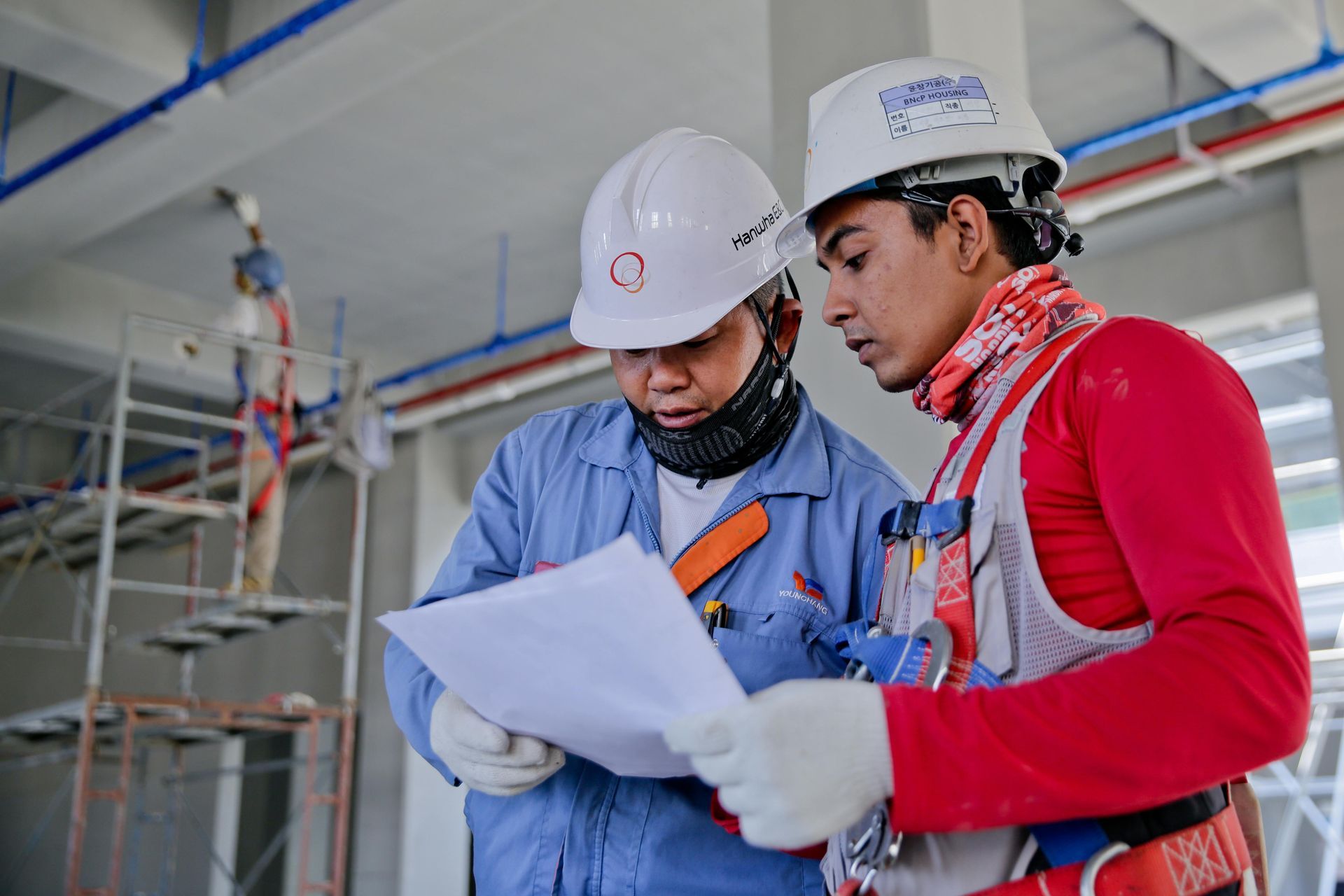 Two construction workers in hard hats and safety gear review blueprints indoors.