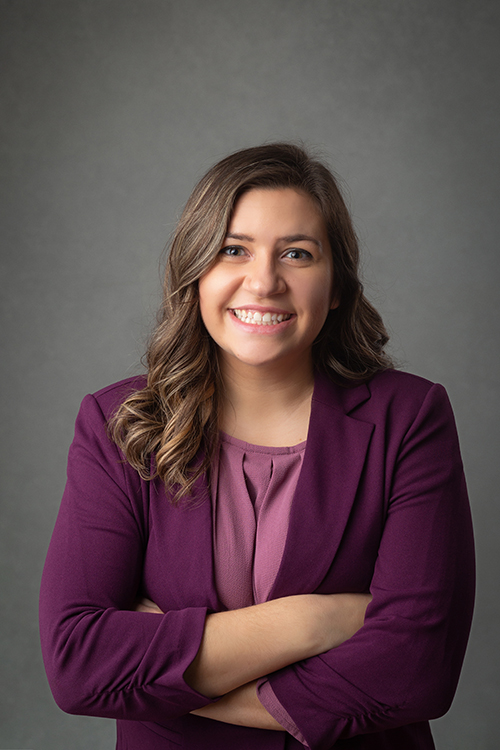 A woman in a purple jacket and pink shirt is smiling with her arms crossed.