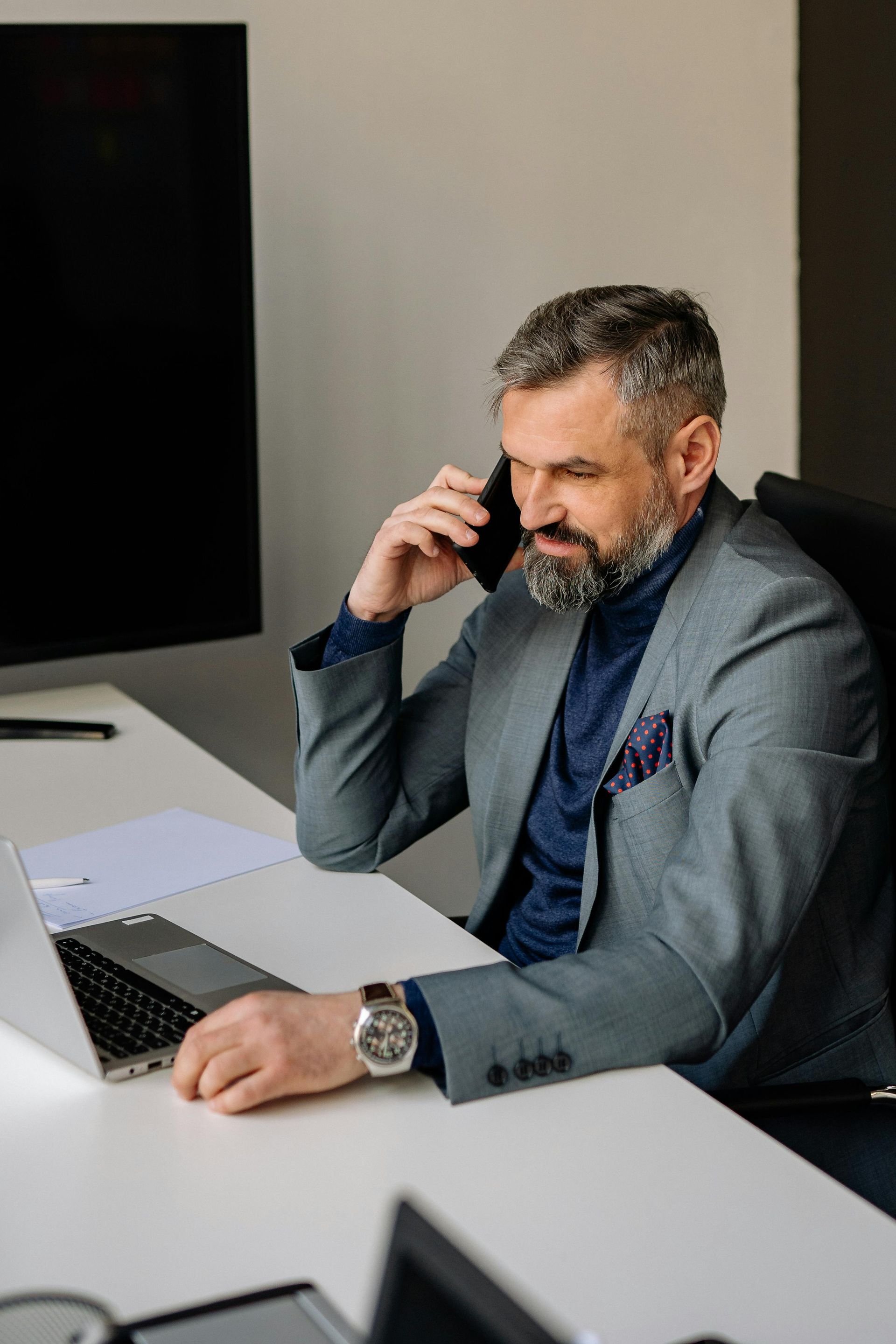 A man is sitting at a desk talking on a cell phone while using a laptop computer.